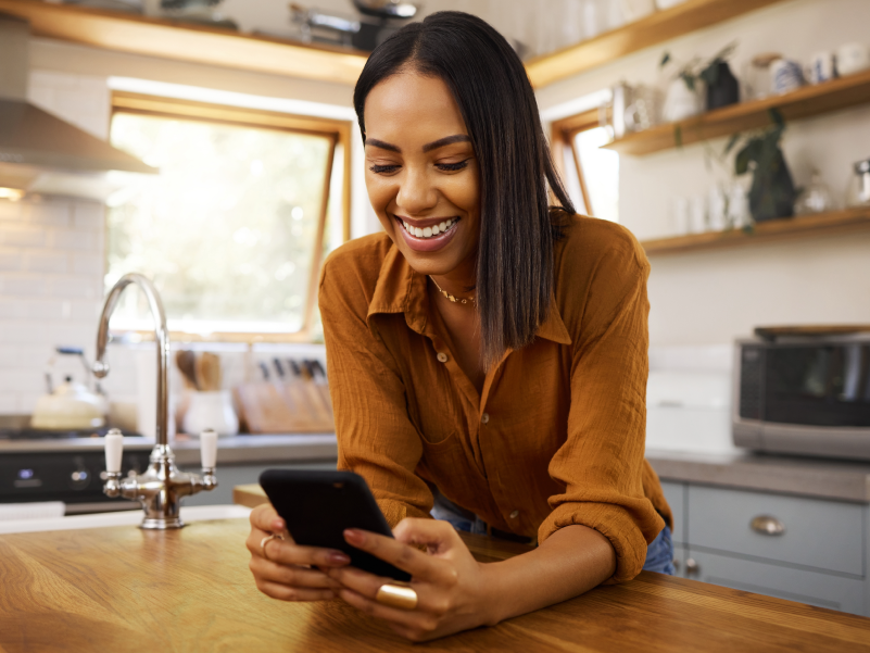 An image of a woman in a kitchen looking at her phone and smiling. 