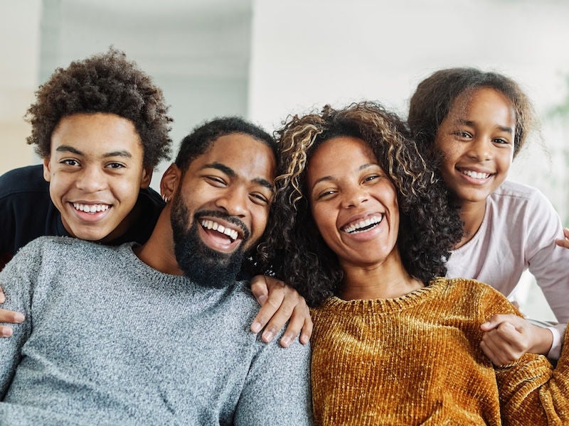 An image of a family with two parents and two teenagers sitting on the couch smiling.